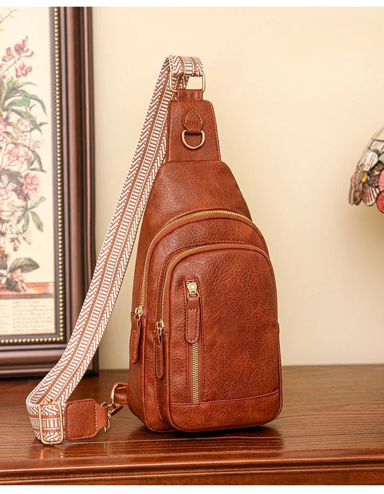 Sac à bandoulière marron en cuir PU avec poches zippées, posé sur une commode en bois, doté d’une sangle textile à motif graphique.
