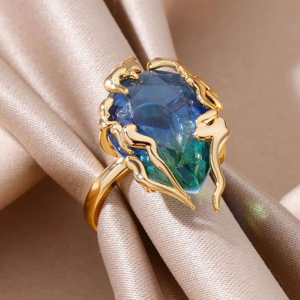 Bague dorée avec pierre zircon bleu et vert en forme de flamme, posée sur un tissu satiné beige noué — ALTAREV