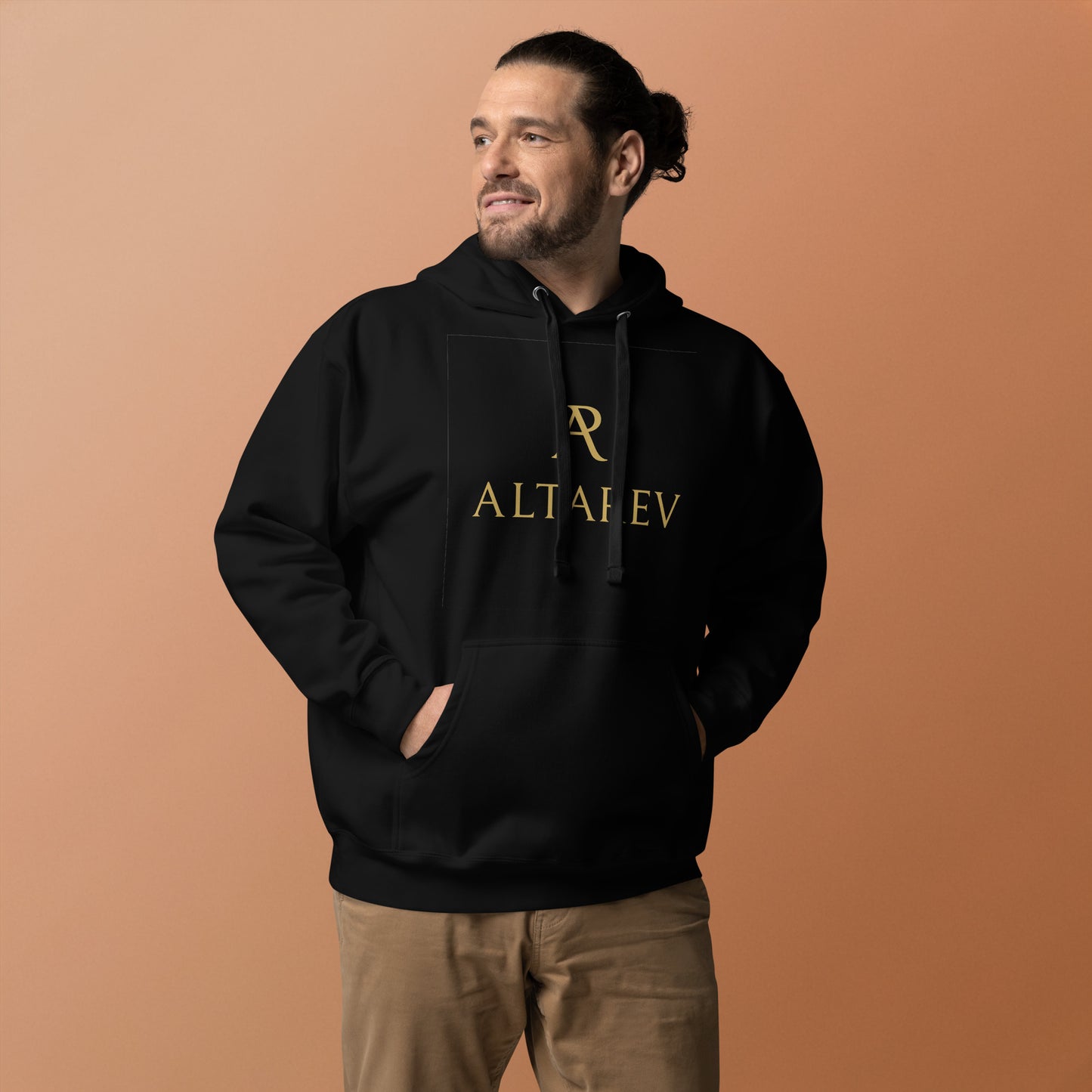 Homme debout portant un hoodie noir ALTAREV avec logo doré, sur fond beige, mains dans les poches, regard confiant.